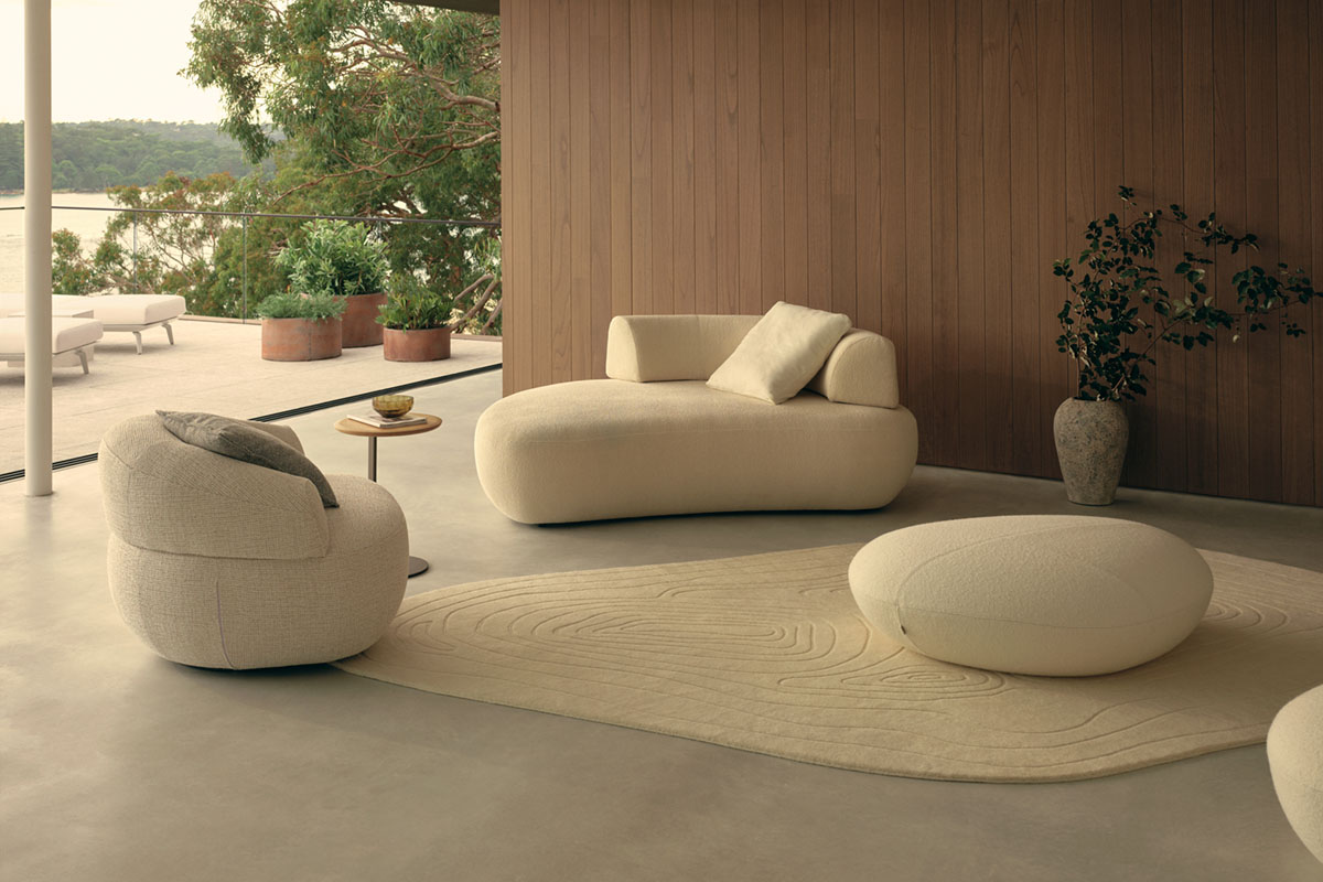 Open-plan living area leading into outdoor area styled with sunlounges. Interior is neutral in colour, spacious and styled with curved furniture.