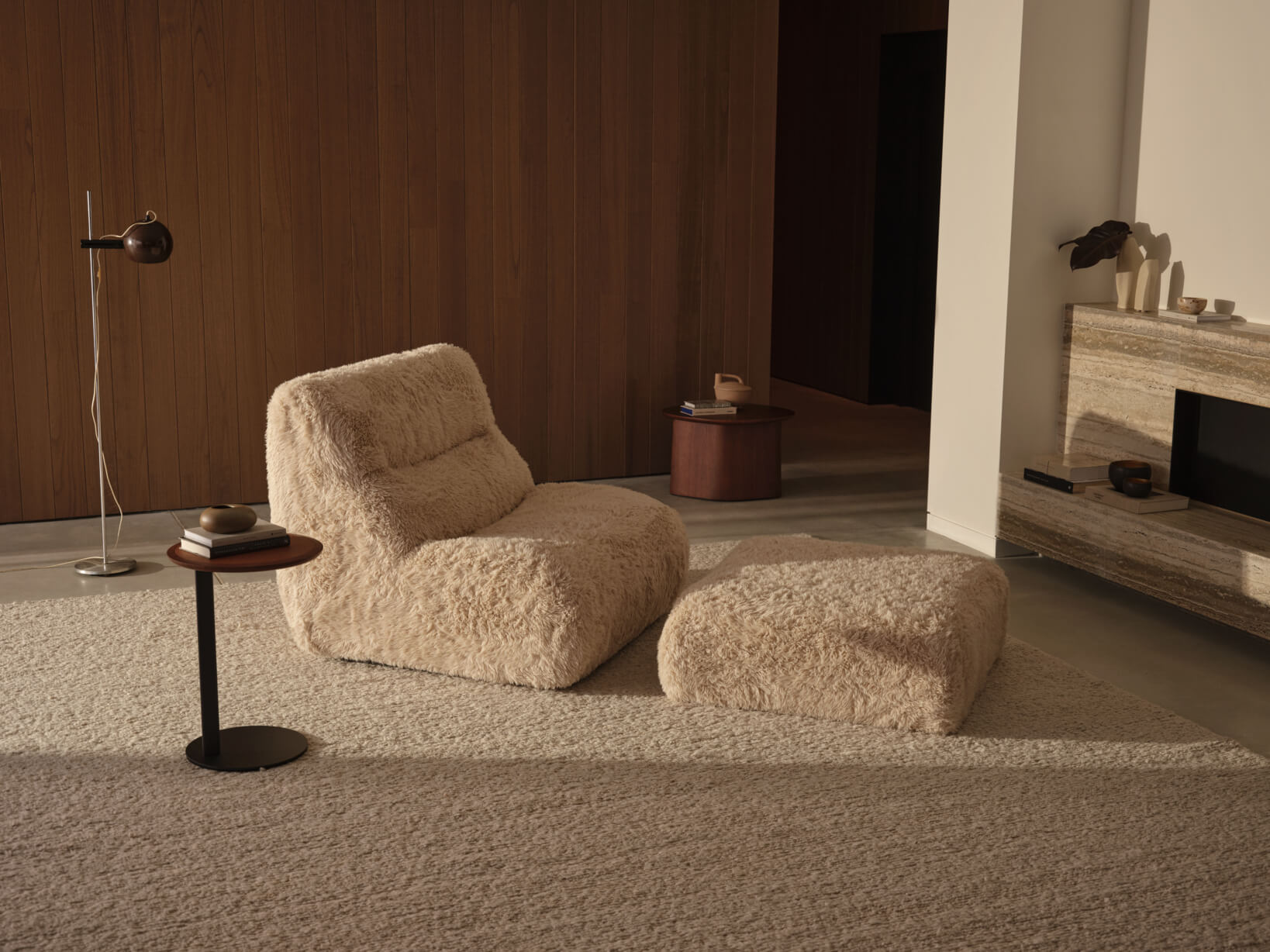 A matching high back sofa chair and ottoman, a perfect reading chair design, sit centrally in a contemporary living room, styled in a faux fur fabric on a soft, warm-toned rug.