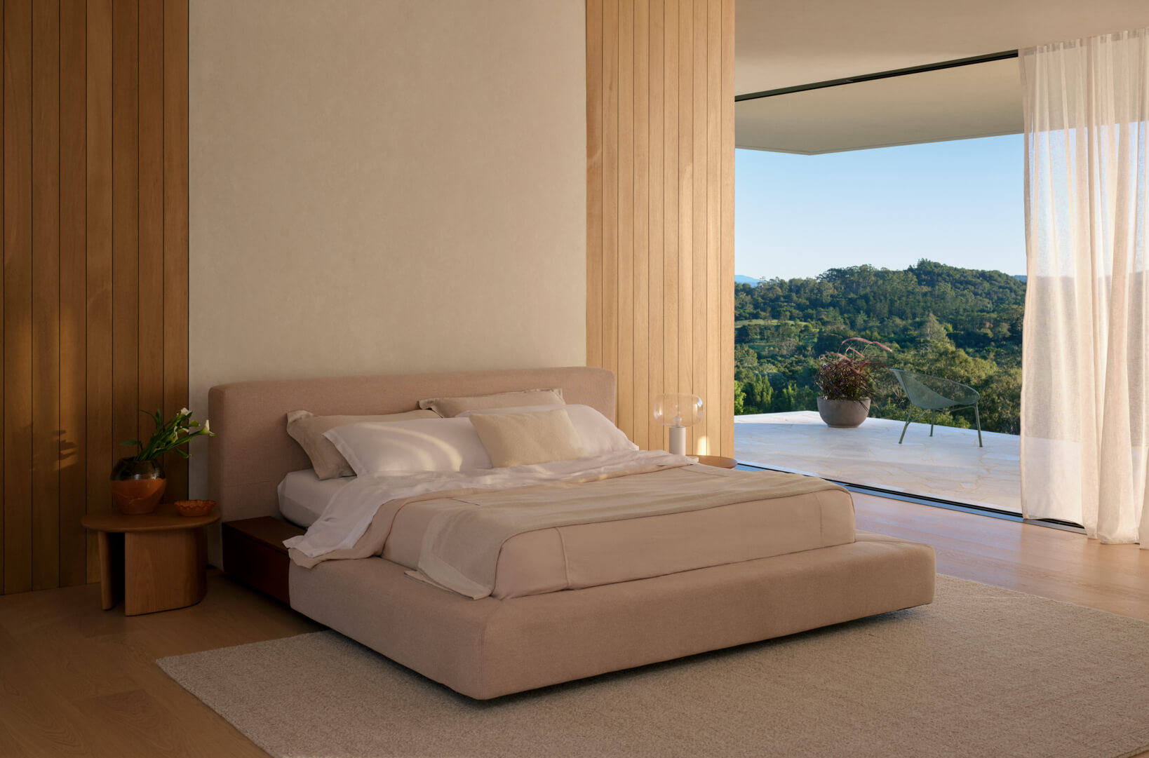 A king size bed in a master bedroom, with an upholstered bedhead in blush fabric, opening to a terrace with landscape views.