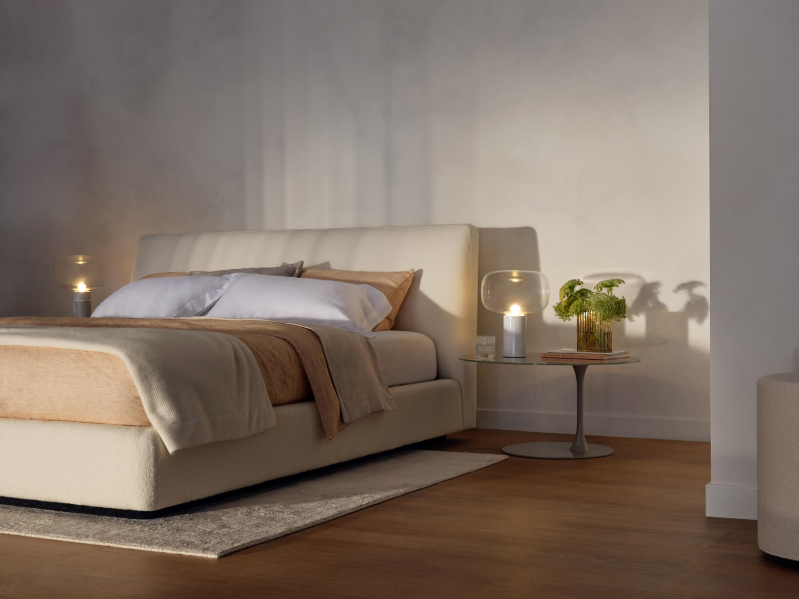 Curved queen size bed with tonal bedding, paired with a round table and ambient lighting in a warm, minimal setting.