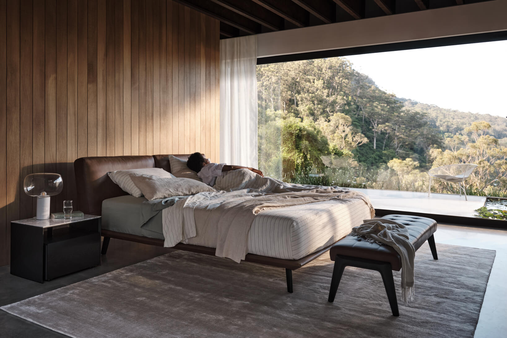 Woman lies on her side in a contemporary bedroom facing a window with expansive hillside views.