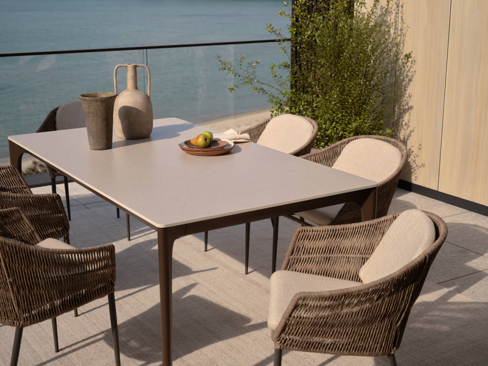 An outdoor dining table with six woven outdoor dining chairs, styled with decorative objects and set beside the ocean.