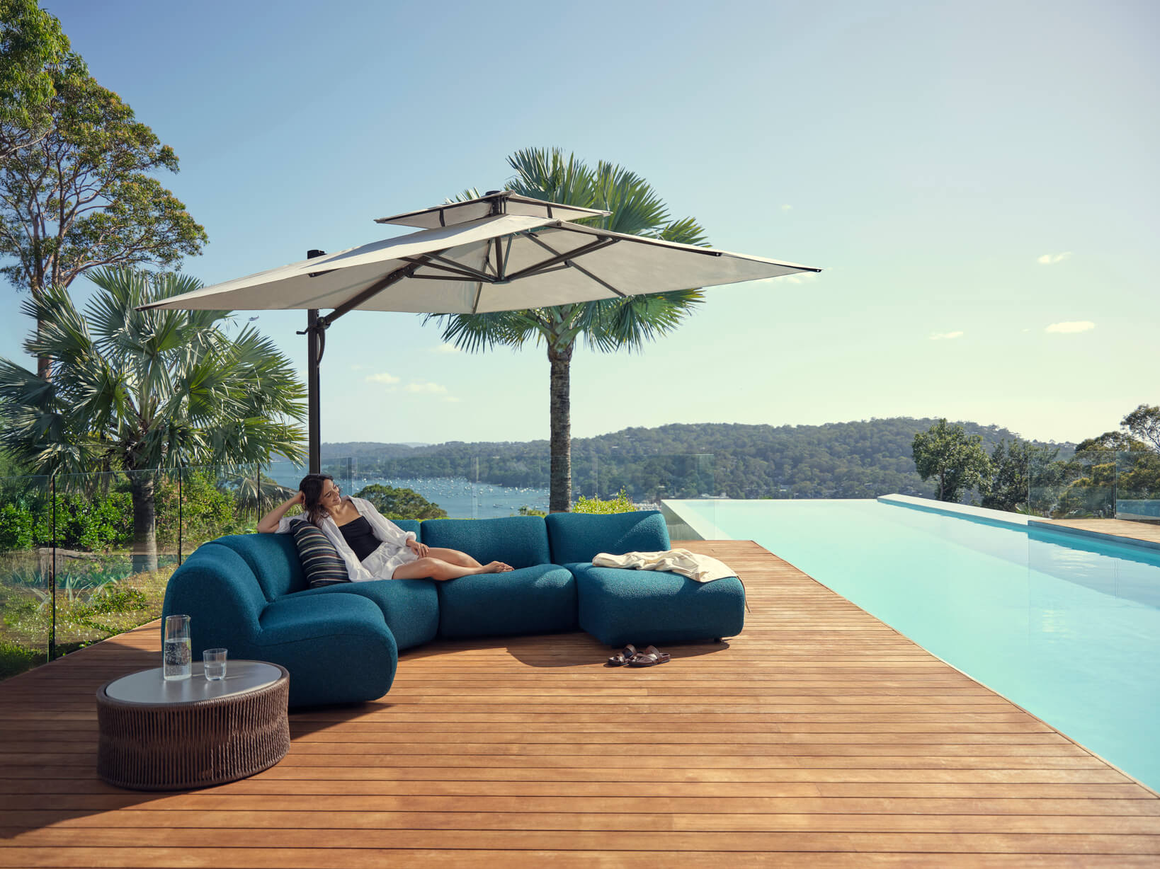 A woman lounges on a modular outdoor sofa styled in a blue bouclé fabric, the setting is shaded by an outdoor umbrella and overlooks a pool and landscapes.
