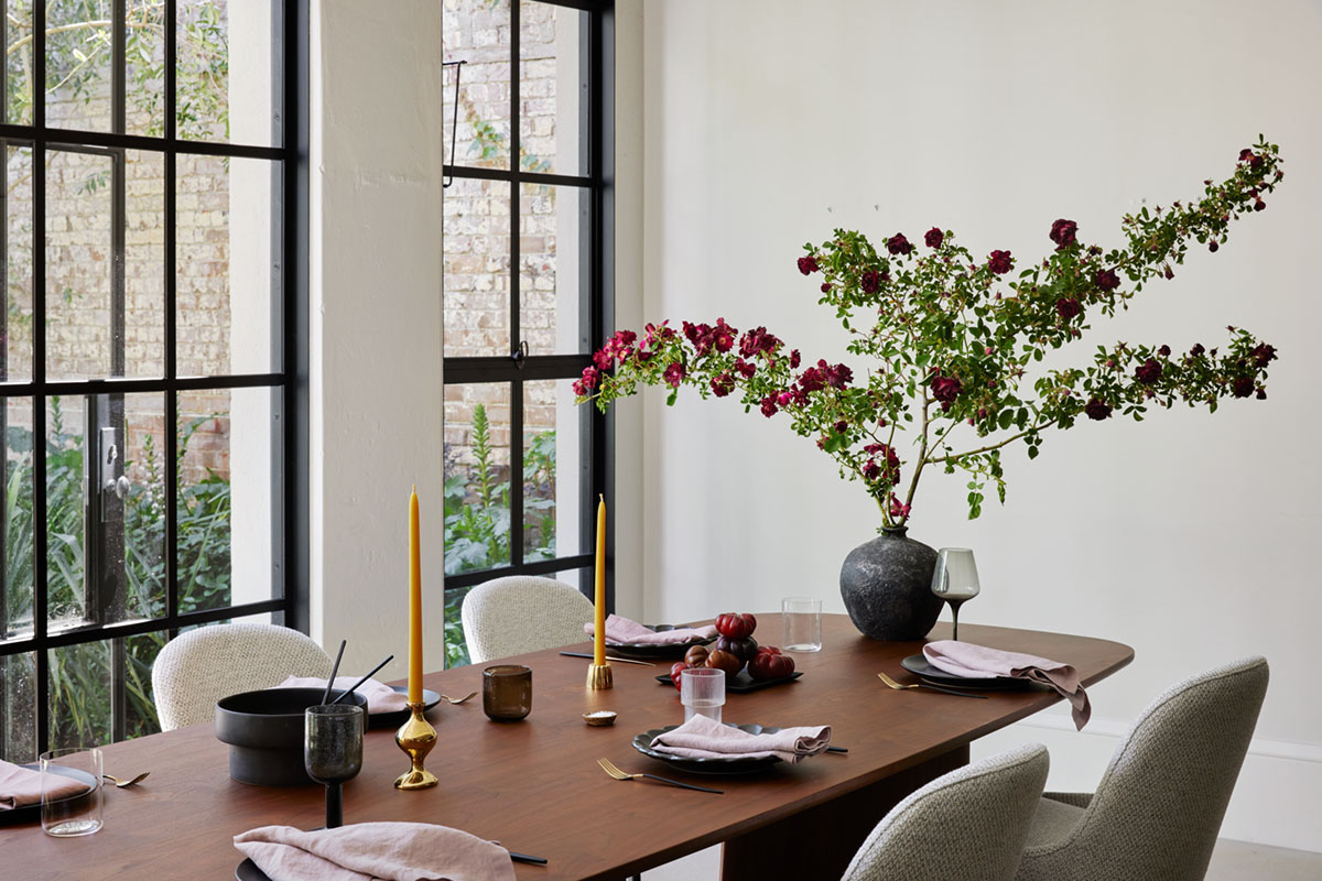 A long oval dining table in a warm walnut timber styled with gold candlesticks, a tall floral arrangement and blush metallic tableware.