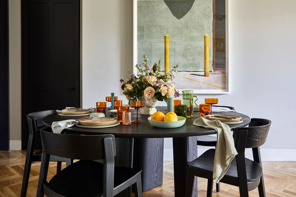 Round dining table styled with colourful fresh flowers, candles and fruit beneath abstract artwork in a modern dining room.