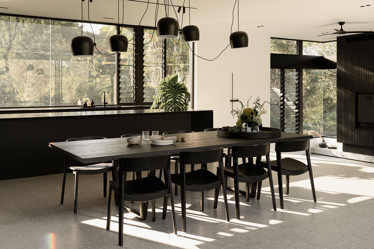 Contemporary open plan dining and kitchen space with black timber table and chairs. Monochrome interior with natural light and greenery.