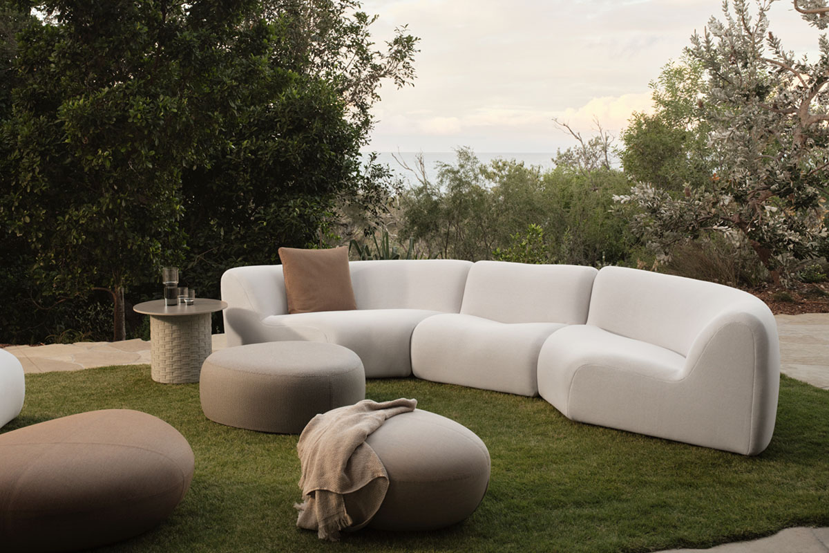 A white bouclé modular outdoor sofa in a grassy garden setting, styled with contemporary outdoor side tables and ottomans.